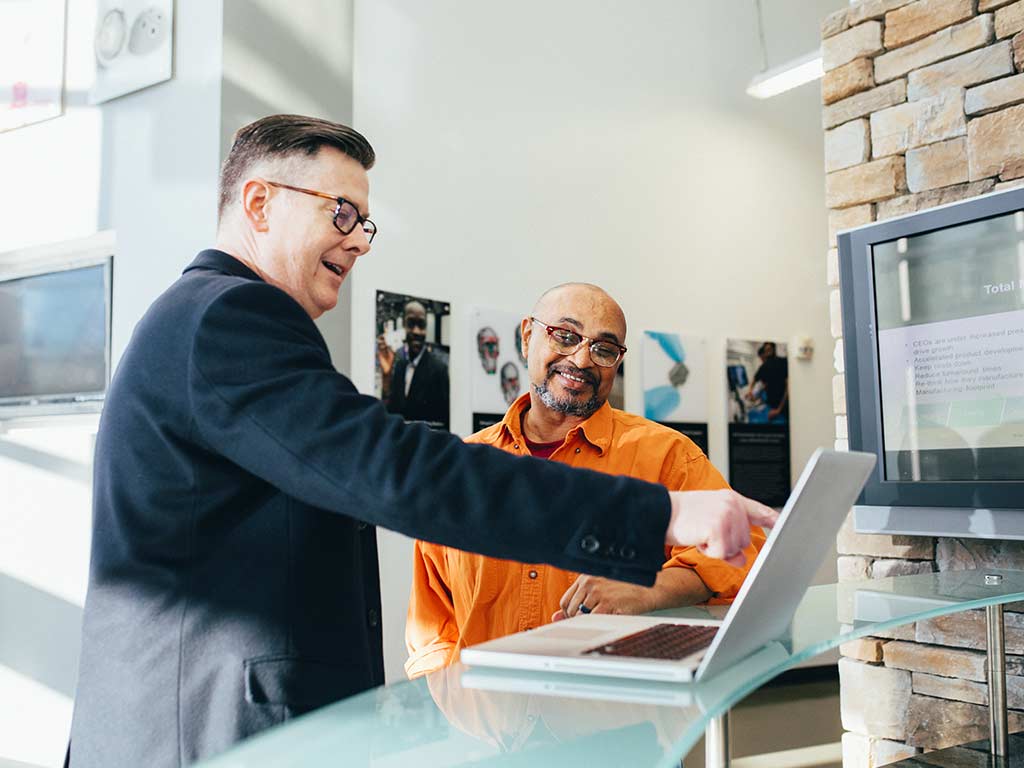 Ein Berater und ein Kunde stehen vor einem Laptop. Der Berater zeigt dem Kunden etwas auf dem Laptop, beide sind glücklich - Startseite